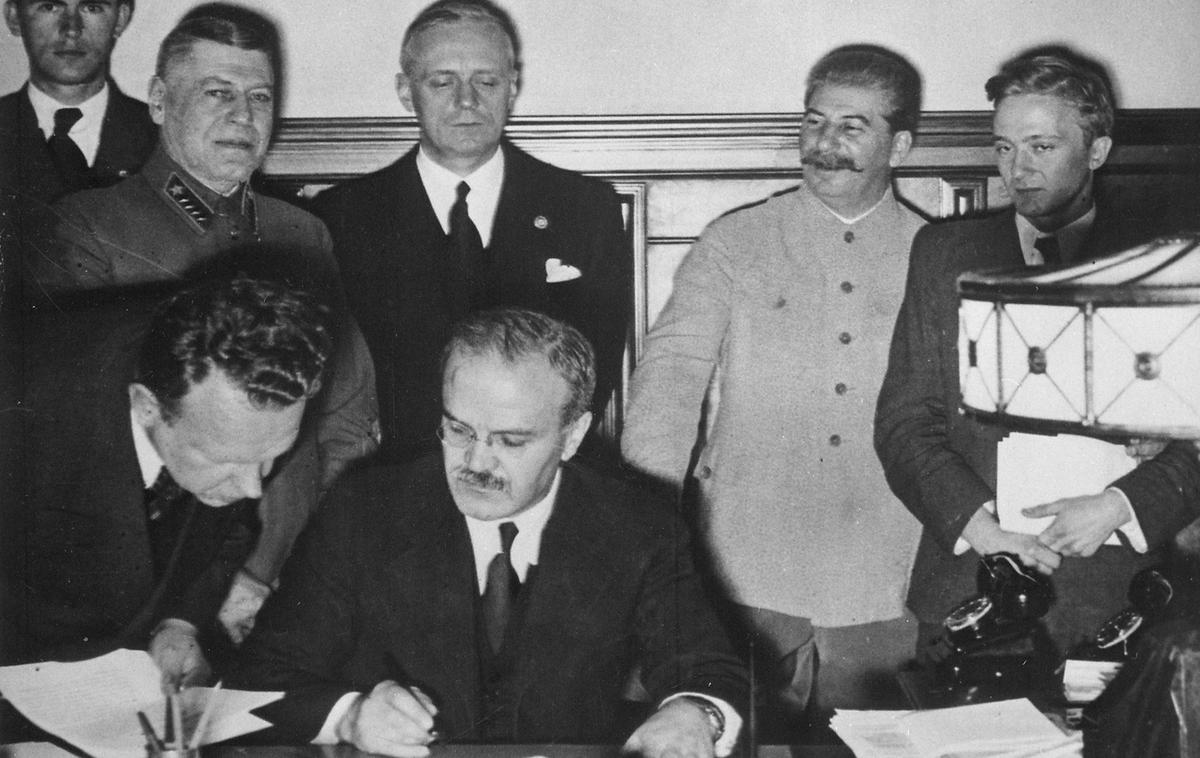 Podpisovanje pakta Ribbentrop-Molotov | Podpis pakta Ribbentrop-Molotov v Moskvi. Sovjetski zunanji minister Vjačeslav Molotov (z očali) sedi za mizo in podpisuje pakt. Za njim stoji nemški zunanji minister Joachim von Ribbentrop. Desno ob njem je Stalin, ki je več kot očitno zelo dobre volje. | Foto Wikimedia Commons